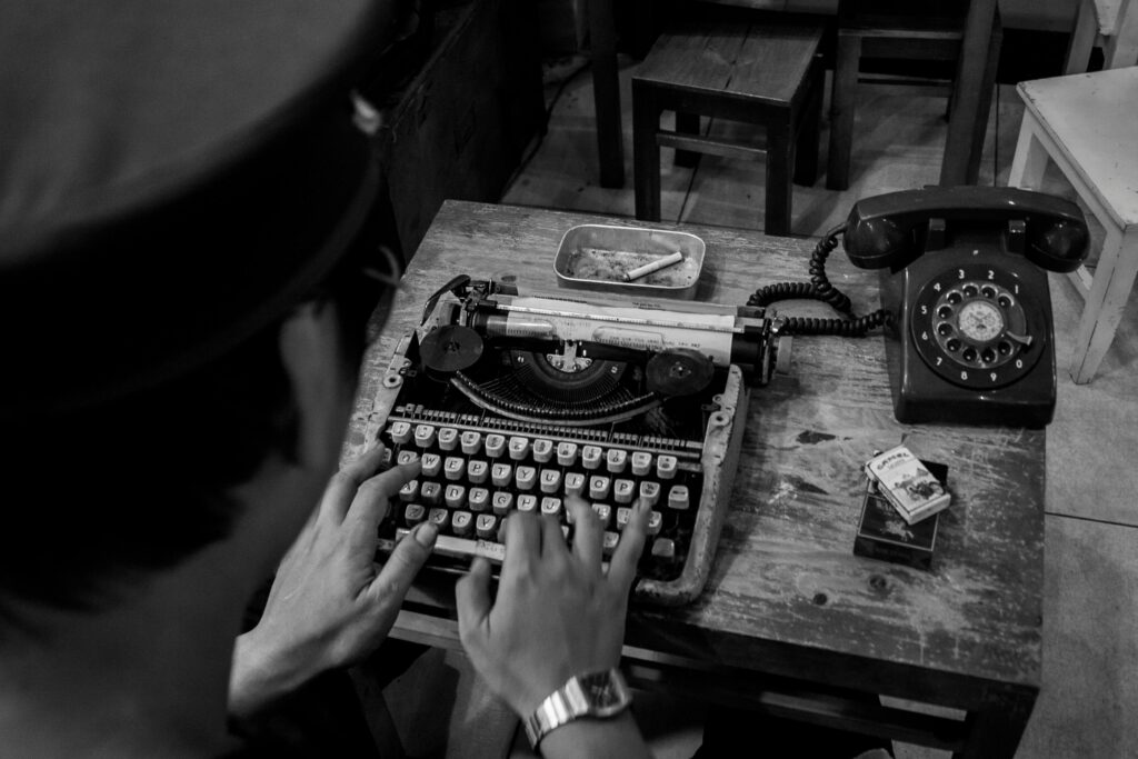 Black and white image of a person typing on a vintage typewriter with a retro telephone nearby.